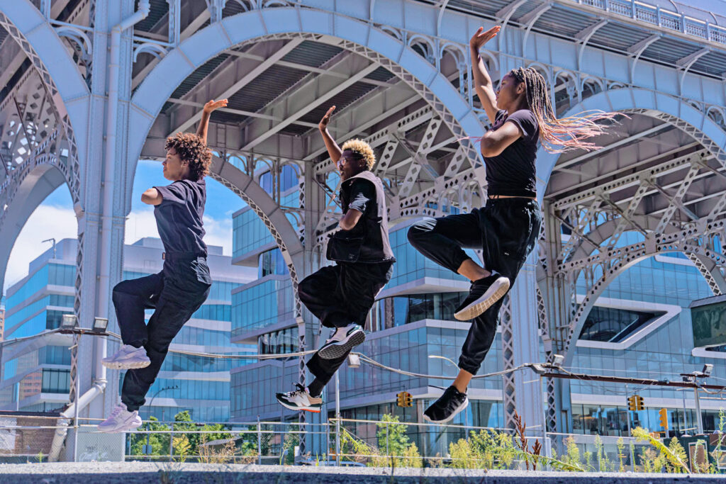 Three dancers wearing black clothes and sneakers are caught midair as one foot kicks up to their opposite knees, one hand upraised and the other pressed to their chest. They are in front of a nearby underpass, scraggly greens and streetlights visible nearby.