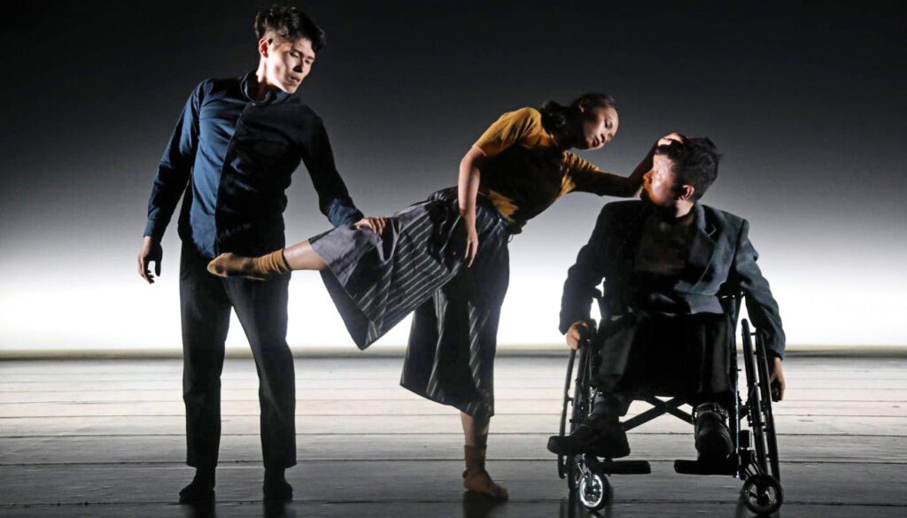 Two standing dancers on the left, one dancer on the right in a wheelchair.