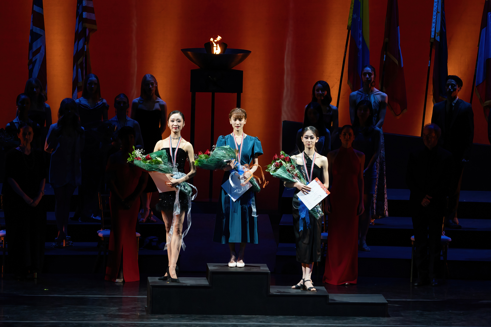 Three young women stand on an Olympic-style podium, wearing formal dresses and high heels and carrying bouquets of flowers. They each wear a medal around their neck.