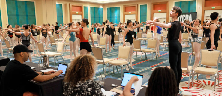 a panel of judges watching students do a ballet barre in a ballroom