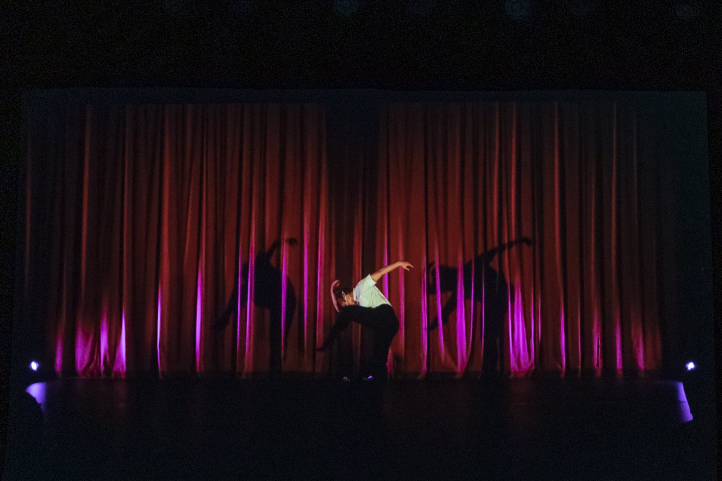 A still from Madeline Maxine-Gorman's "Between Myself" shows her in front of a large red curtain with pink lighting, dancing in a white button-up shirt and black slacks.