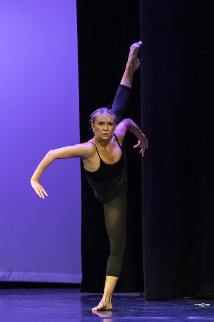 Quinn Starner performs a contemporary solo onstage in a black leotard and black tights. She does an extreme tilt with her arms flexed and inverted inward.