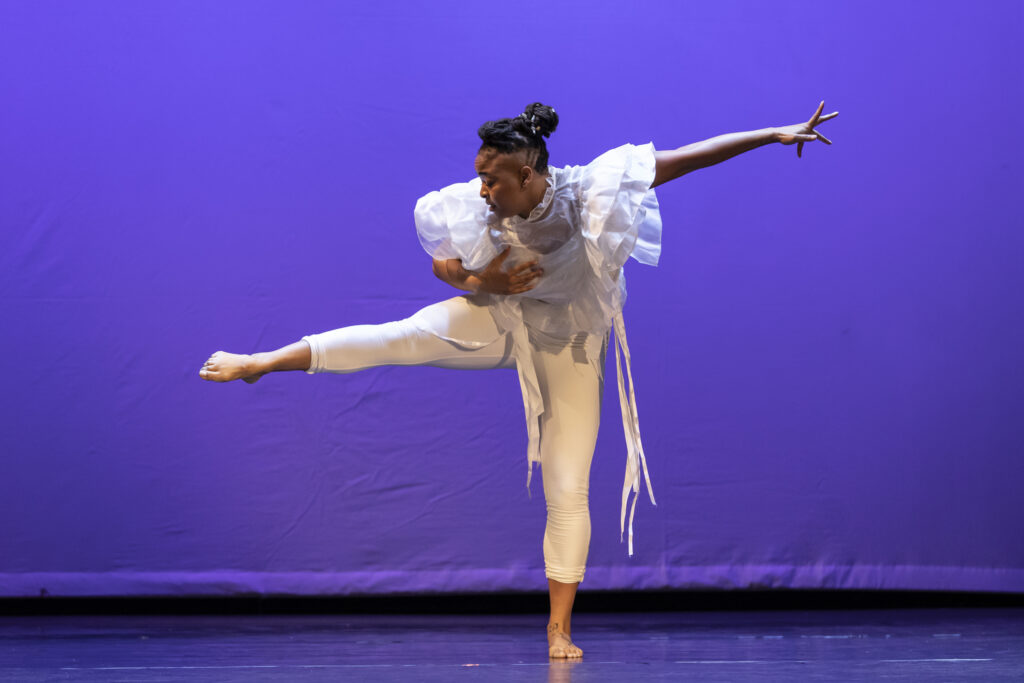 Tendayi Kuumba performs a solo in a white blouse and white bodysuit. She balances on her left leg, her right lifted in a parallel attitude to the side. Her arms mirror her legs.