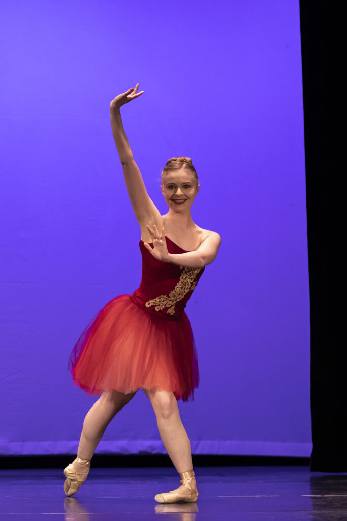 Erin Casale performs a variation from "Walpurgisnacht." She wears a red and gold costume with pink tights and pointe shoes. She poses in a playful lunge forward, her back leg in tendu and her arms flexed, one above her head and one in front of her chest.