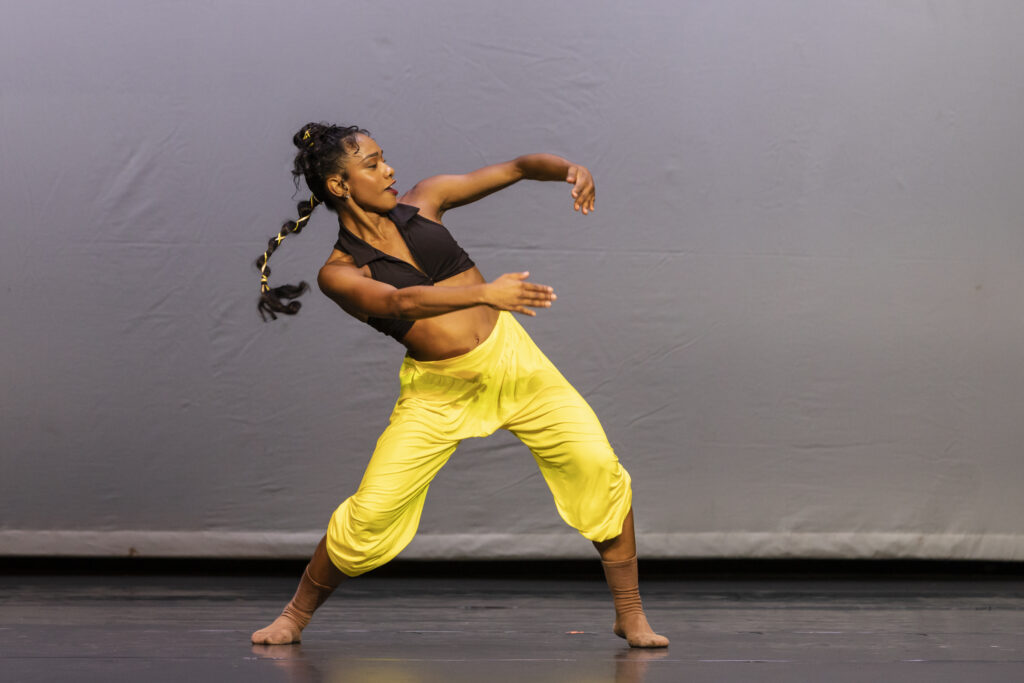 Dandara Veiga dances a high-energy solo in a black crop top and bright yellow culottes, her long black hair in a high ponytail with a yellow ribbon weaved through it. She shifts her weight with her legs bent and feet wide, arms in a wide first position as she bends to the side and smiles.