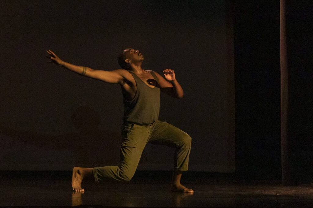Jordan Demetrius Lloyd performs a solo in dark lighting and loose brown and gray clothing. He lunges on his right leg, chest turned upward and right arm lifting up and back as his left arm curls into his chest.