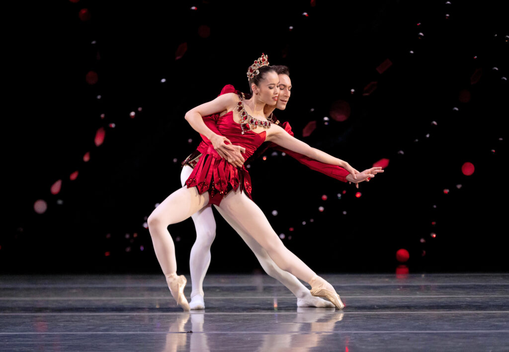 A female dancer lunges to the side en pointe, supported by her partner standing behind her, his arm around her waist as he matches her angular pose. Both look towards their entwined arms as they extend on a downward diagonal. They are cheek-to-cheek. She wars a ruby red short dress, pink tights, and pointe shoes. He wears white tights and a tunic in a matching ruby red.