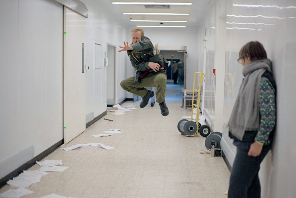 a woman leaning against the wall in a hallway watching a male actor jump in the air 