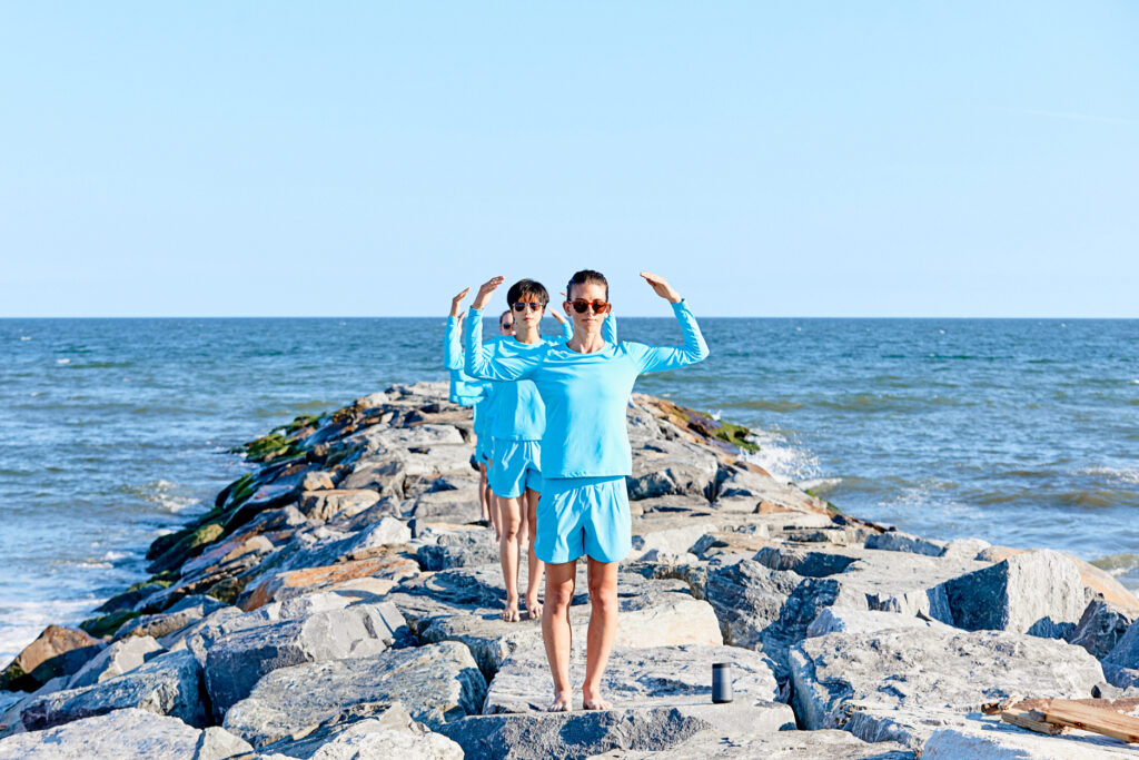 On a rocky outcropping leading to the see, a single-file line of dancers in cerulean blue stand facing the camera. They stand with their legs in parallel, arms raised to shoulder height and bent at the shoulder so their fingertips arc toward their heads. The sky is cloudless and pale blue.