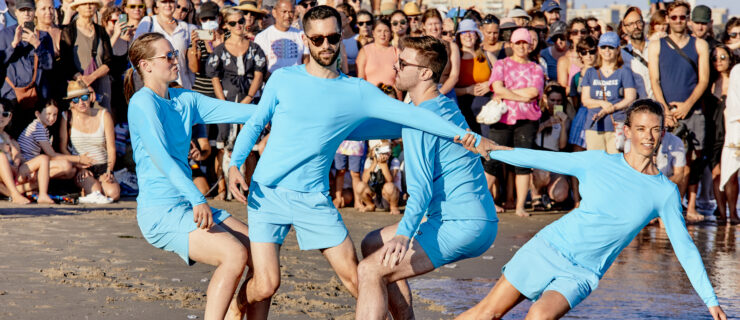 Four dancers in matching cerulean blue costumes in an interlinked counterbalance on the wet sand of a beach. A crowd behind them watches.