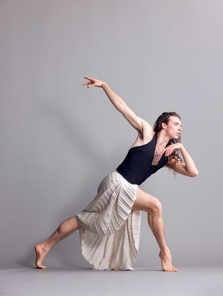 Aaron Choate poses in a forced arch lunge, long brain hair tumbling down their back as a white skirt drapes over their legs. One hand is tucked under their chin, the other extended elegantly behind them.