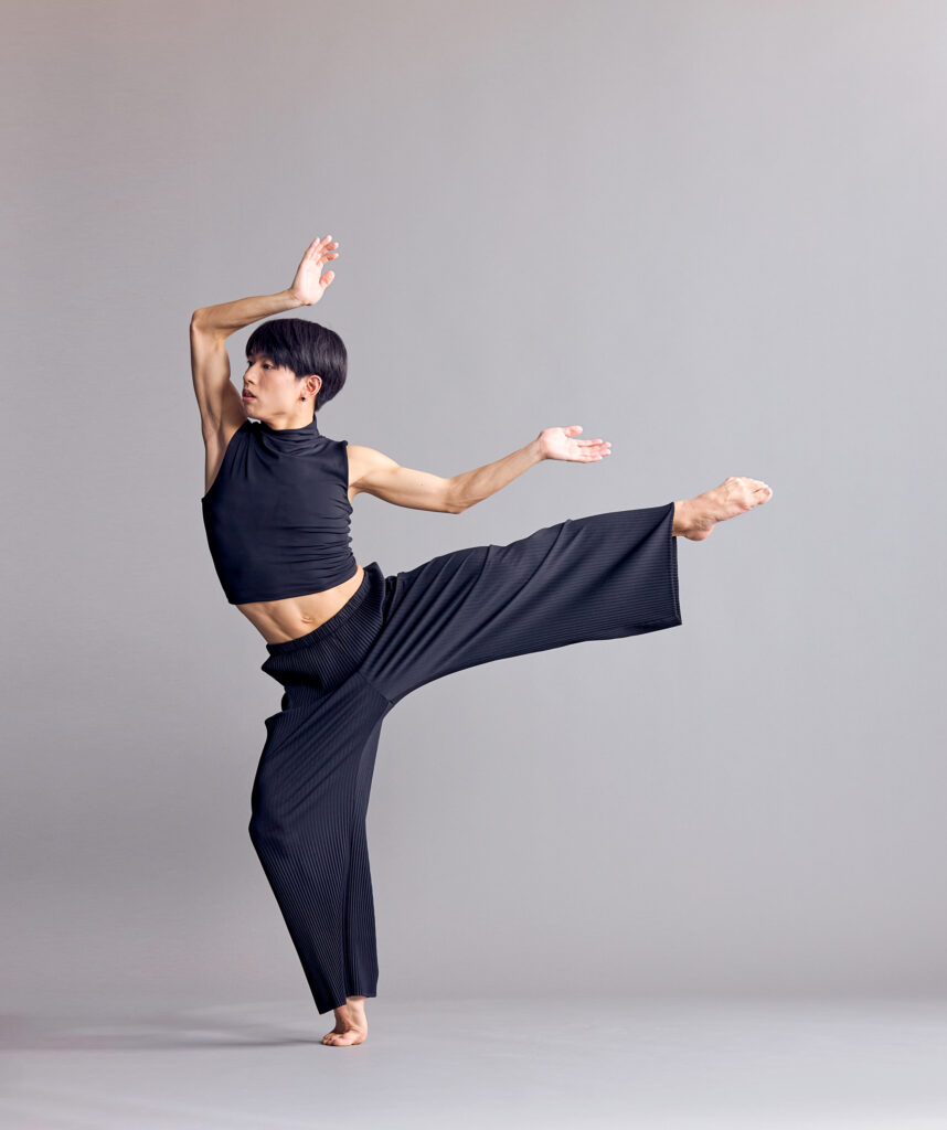 Shota Miyoshi balances in a forced arch back attitude. His working side arm is palm up, elbow bent as it extends back; the other hovers over his head. He wears a black crop top and wide-legged black trousers.
