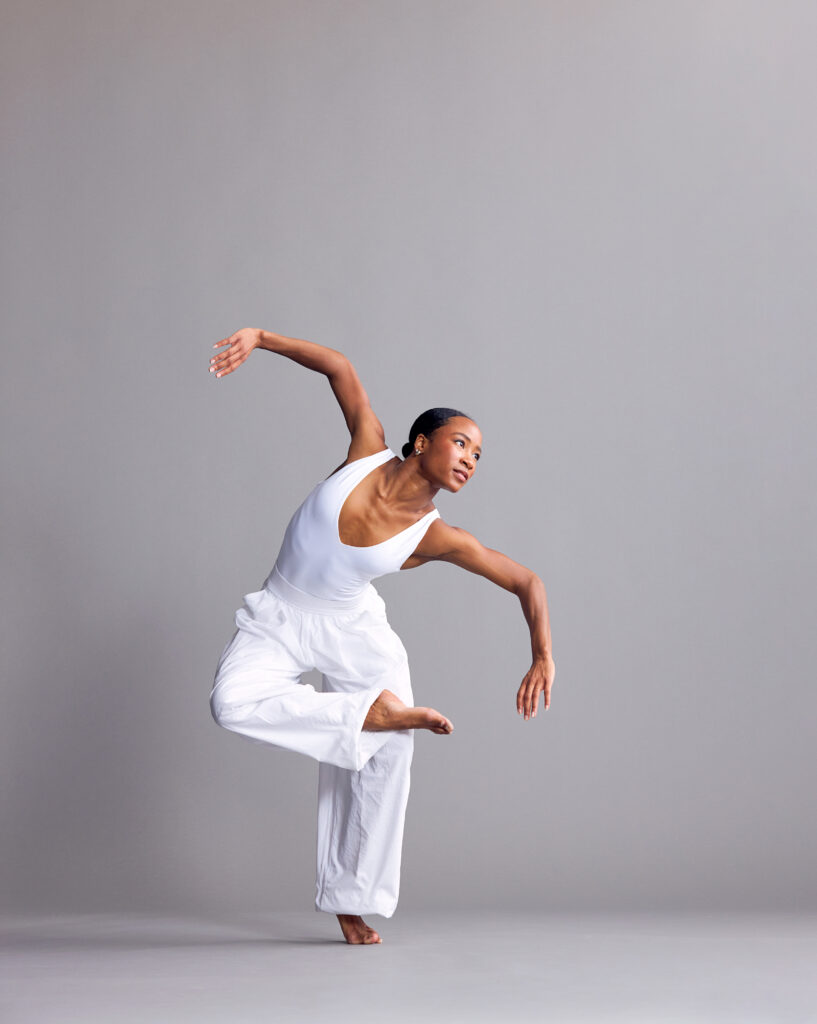 Alexandria Best poses in a white jumpsuit. One foot crosses over the opposite knee, curving arms raised to shoulder height as she leans over her supporting leg. She gazes off-camera, past her lower shoulder.