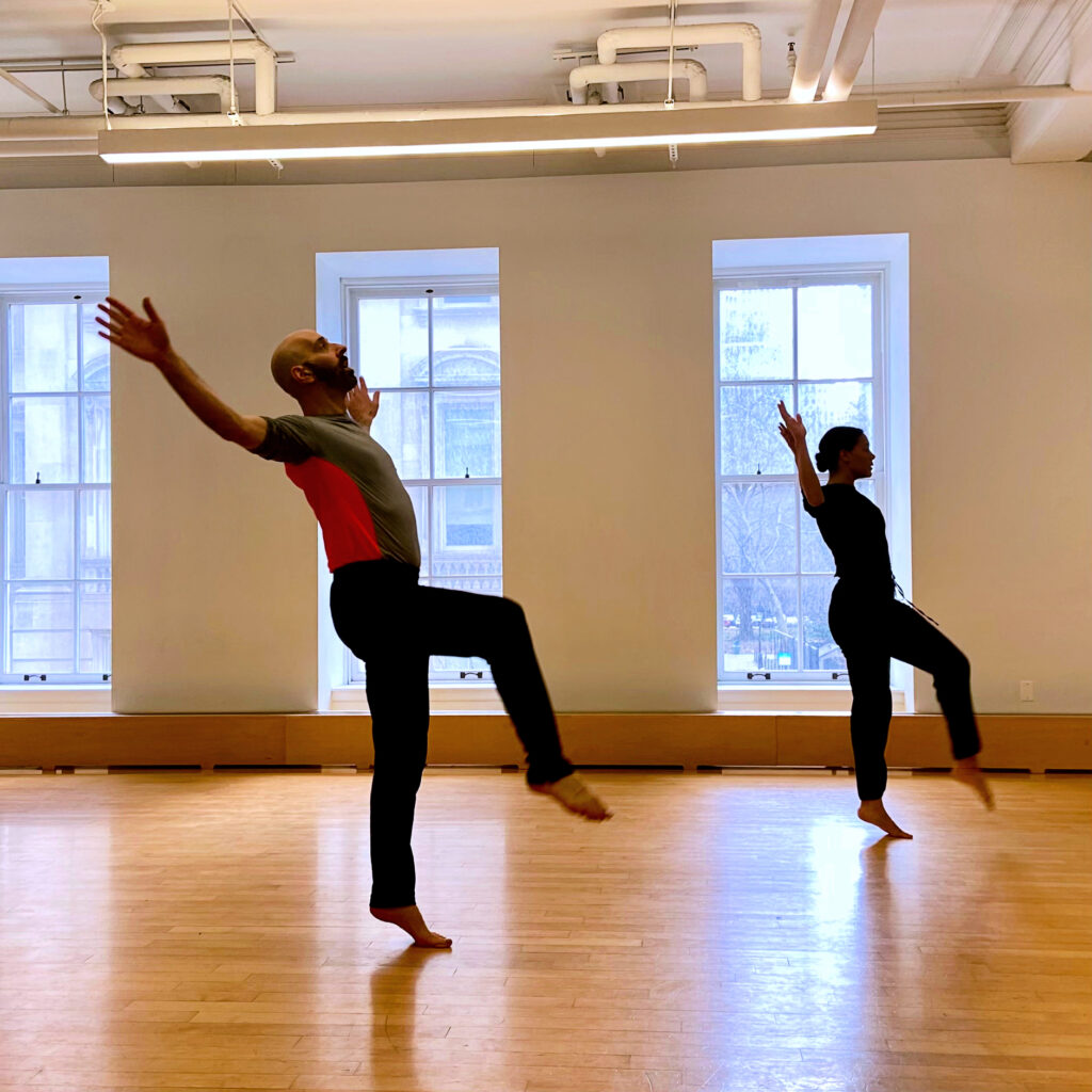 two dancer balancing on one leg with arms out in a studio with large windows