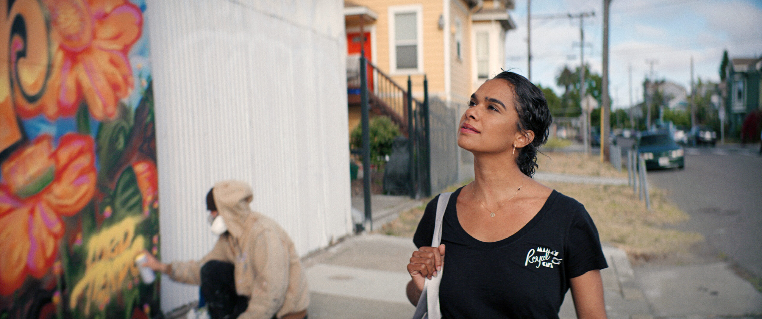 In a film still, Copeland, a light brown-skinned Black woman wearing a black t-shirt, walks along a city street, admiring a painted mural of flowers.
