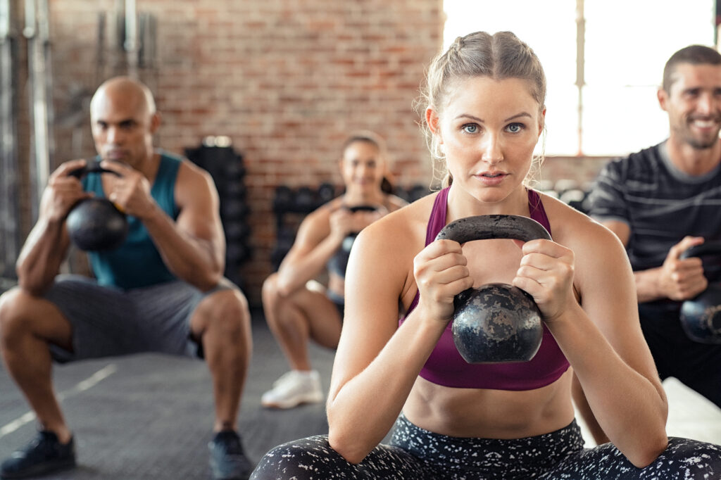 Group of fit people holding kettle bell during squatting exercise at cross training gym. Fitness girl and men lifting kettlebell during strength training exercising. Group of young people doing squat with kettle bell.