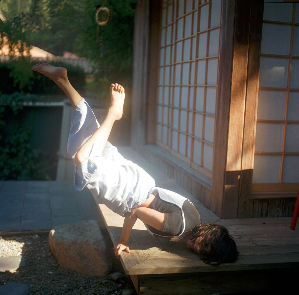 A dancer in baggy white overalls poses on a narrow, sun-drenched porch. Their legs bend their feet toward the sky behind them as they hug the floor, elbows bent and palms pressing down to give them leverage.