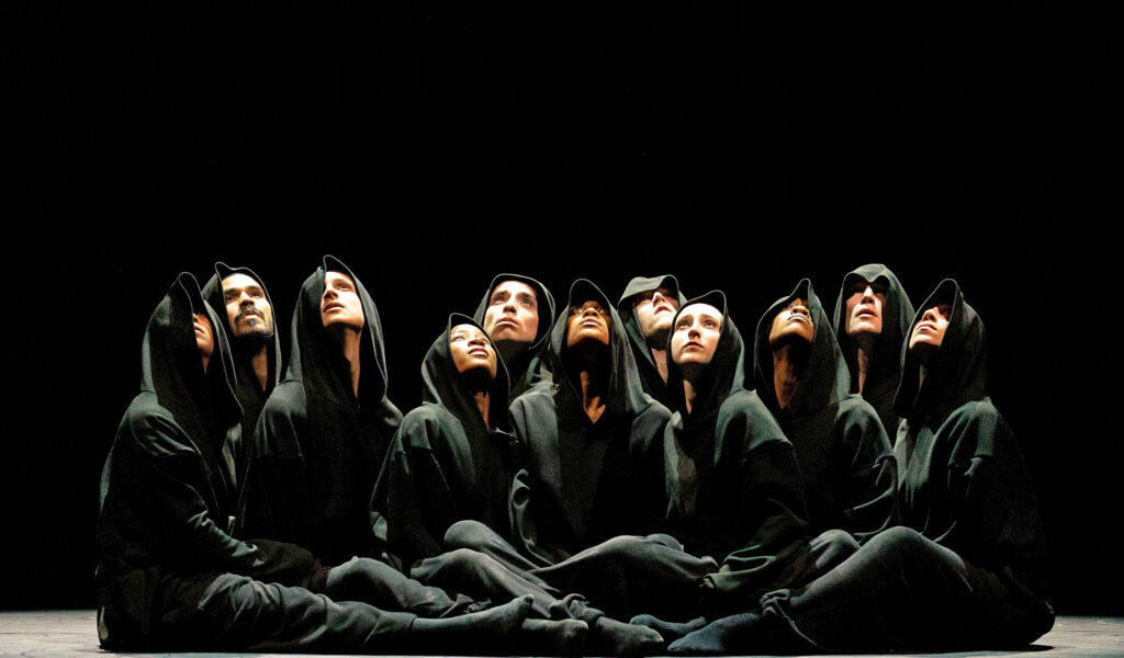 Eleven dancers sit clustered tightly together, their upturned faces peering out from beneath obscuring black hoods.