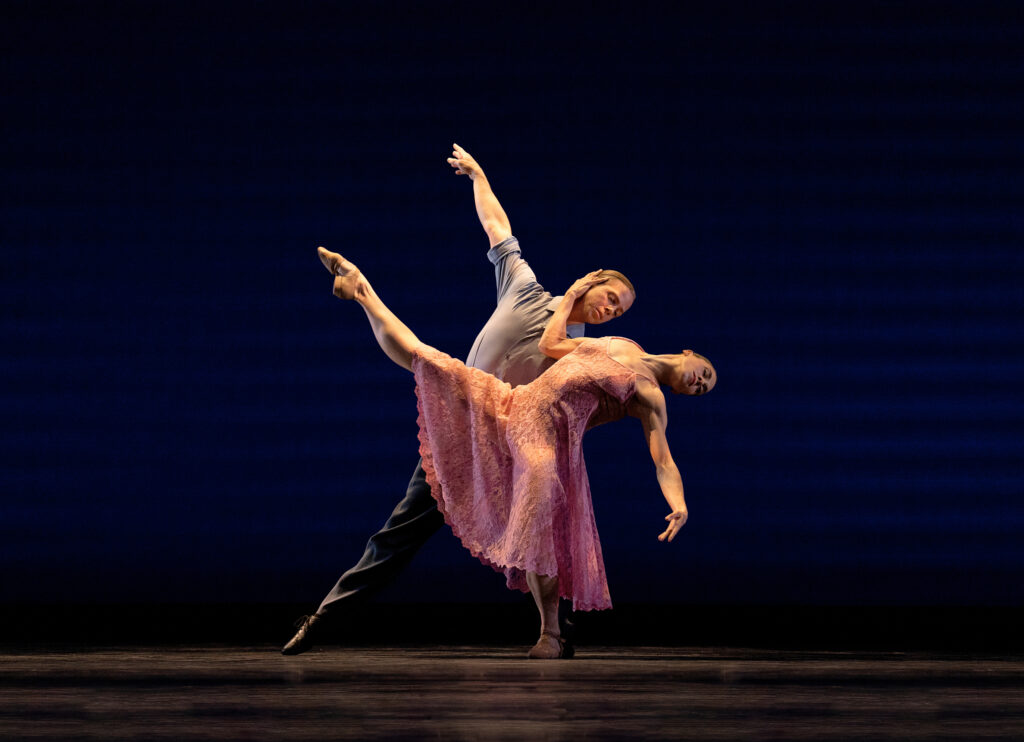 A dancer in a long pink dress extends their leg forward as she arches back, head and arm languidly dripping towards the floor. Her partner supports her with one arm around her waist, free arm extended to match her leg. The first dancer's upper arm cradles her partner's face.