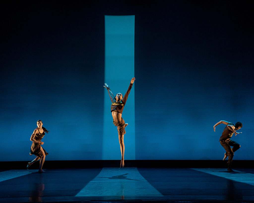 Three dancers on a blue-washed stage. The one at center is in a column of lighter blue light as they jump, arms upraised as one leg extends back. On either side and a bit upstage, two dancers are caught mid-run facing stage left.