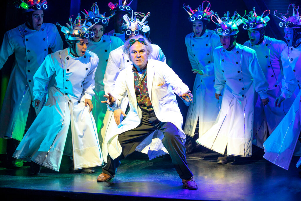 Doc Brown is surrounded by ensemble members wearing exaggerated versions of his white lab coat and goggles. The ensemble leans towards him as they sing, their headpieces recalling quintessential mad scientist villains. Doc Brown is in a dynamic stance, a look of concern on his face as his hands rise.