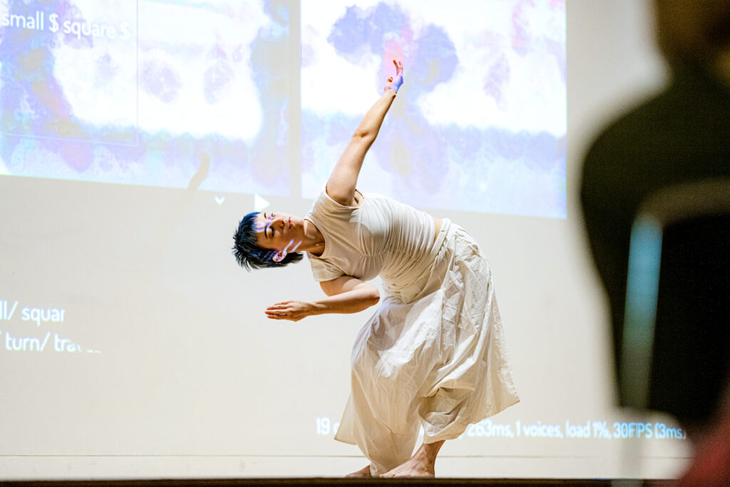 a dancer wearing all white leaning laterally with one arm extended up, a screen projection is behind her