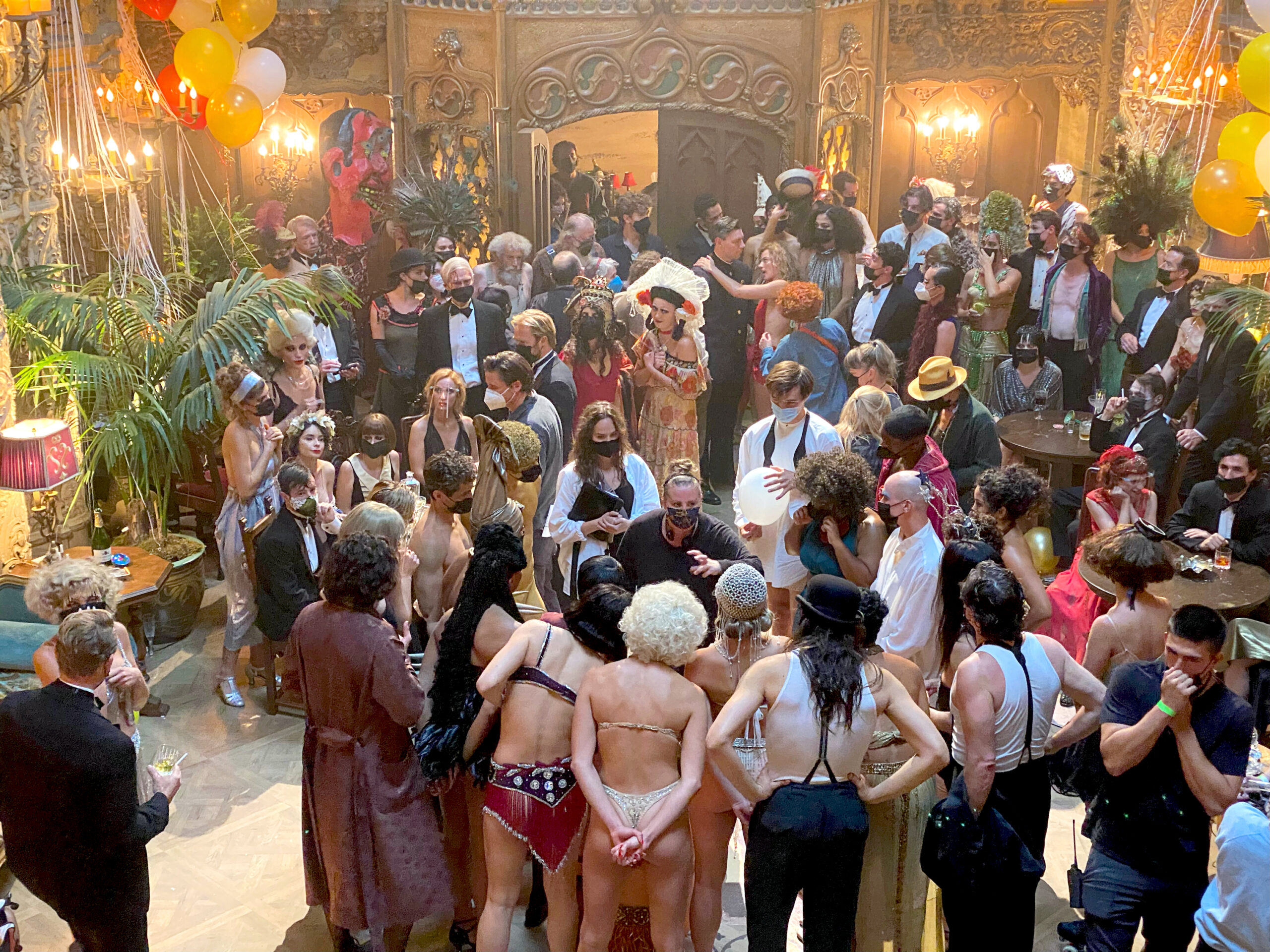 An aerial view of an opulent film set in between takes. Claire Elizabeth Ross and Mandy Moore both wear face masks as Moore speaks at the center of a circle of costumed performers. Several dozen actors and crew members are arrayed around the set.