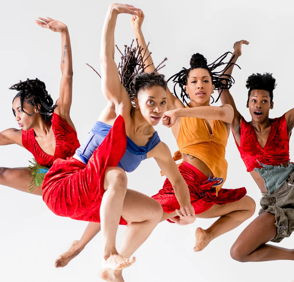Four dancers are captured midair, legs pulled up beneath them and arms outflung in different positions. Each wears either a red satin crop top or a skirt in the same fabric. Braids fly into the air with the motion.