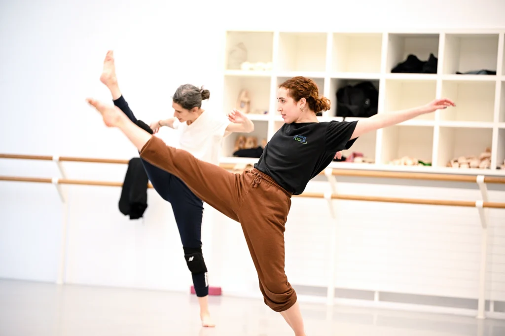 A barefoot dancer extends a pointed foot forward, supporting leg bent as her hips pull her off center. Her arms are flung back behind her shoulders, but her chin is level to the floor, gaze fixed intensely forward past her extended foot. She wears sweat pants cuffed to her calves and a t-shirt.