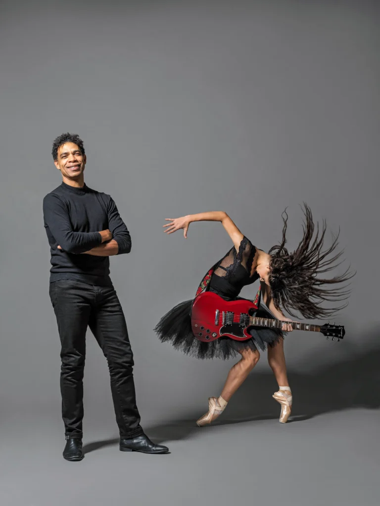 A dancer in a forced arch fourth position on pointe holds a red guitar. Her head is ducked forward, hair flying, as though she headbanged into strumming a chord on the guitar. Carlos Acosta stands smiling, his arms crossed, beside her.