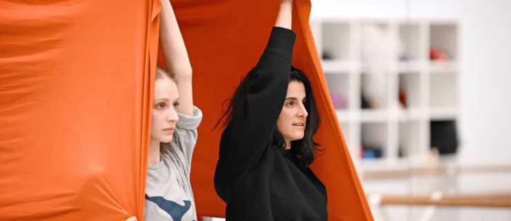Andrea Miller stands beneath a large stretch of orange fabric. She uses one arm to hold it up over her head, the other to pull it down by her hip. A dancer standing beside her beneath the fabric mirrors the gesture.