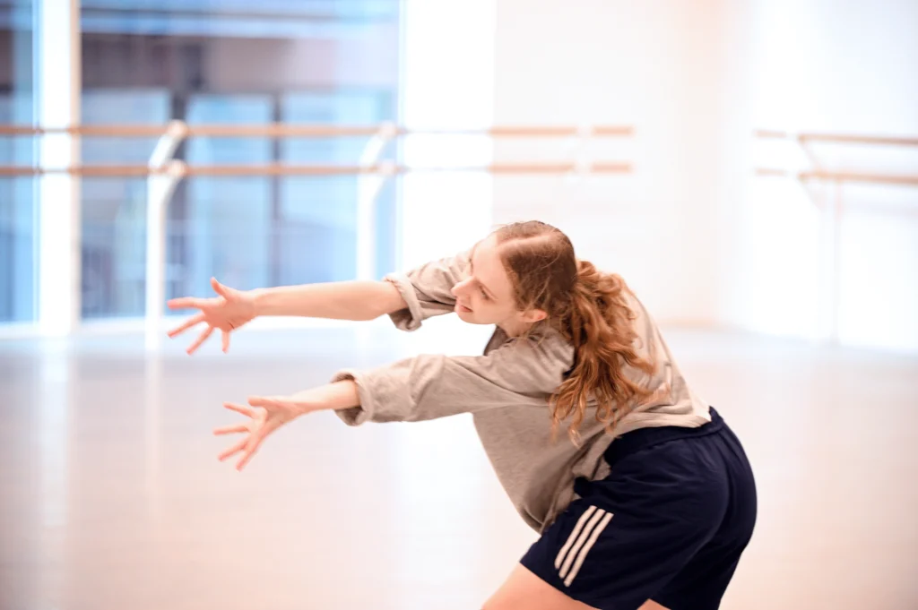 With her back to the camera, a dancer twists and bends to the left, hands splayed wide as she reaches forward. Her knees bend as her hips pull in opposition to where she is reaching. She is in a brightly lit ballet studio; her long hair is in a low ponytail and she wears a baggy shirt and athletic shorts.