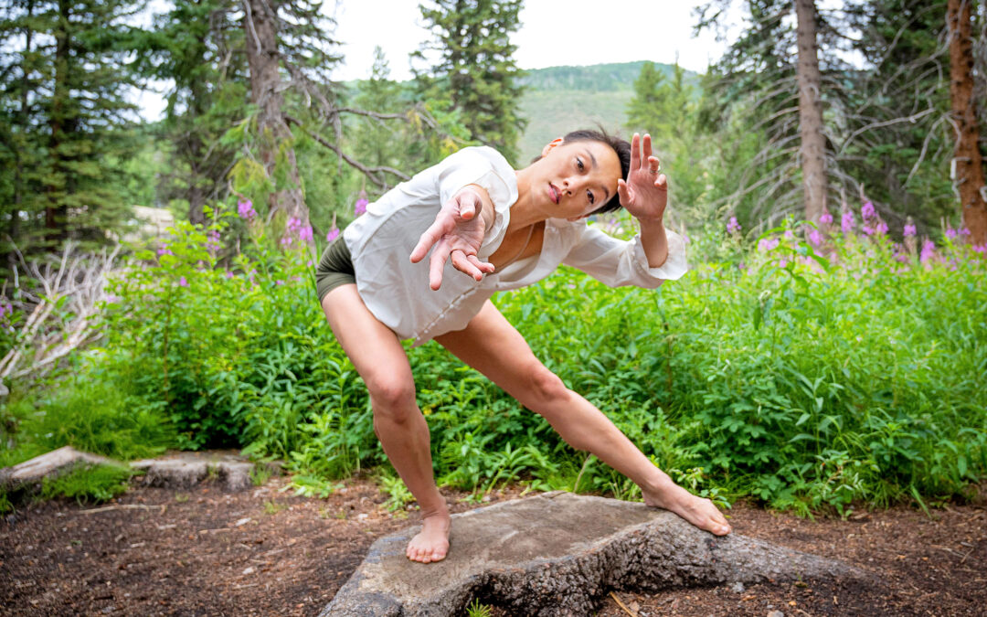 a female dancer standing on a stump in the woods reaching towards the camera