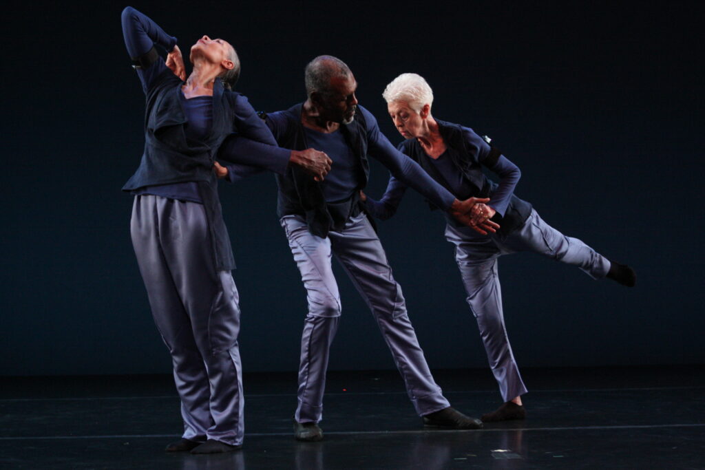 Three older dancers dressed in blue connect. Carmen stands with her feet together, face arching to the sky. She has a hand pressed to the small of her back, and Gus links his elbow through hers as he leans toward her. He looks back to Valda, who places one hand on his hip and the other in his outstretched hand as she steps into a low arabesque.
