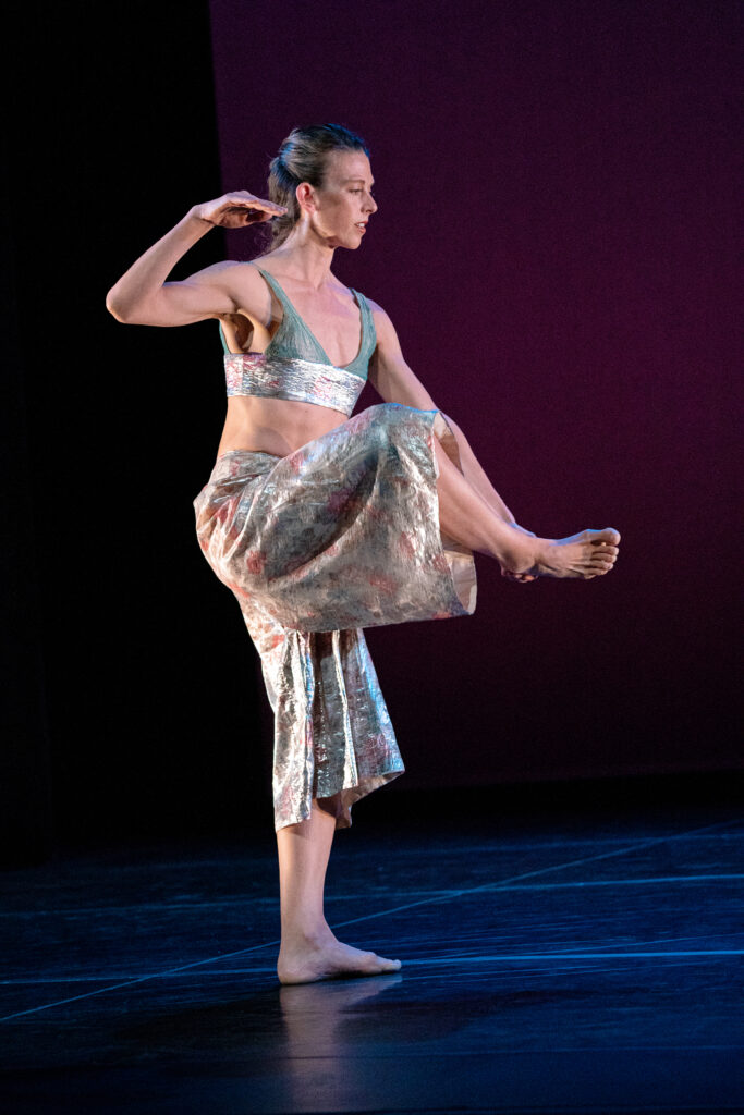 a female dancer wearing wide pants and V-neck bra holding on foot in front of her on stage