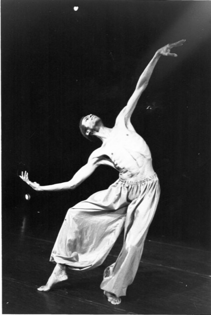 In a black and white archival photo, Gus Solomons jr's long limbs unfurl. His arms curve elegantly towards the sky as he leans to one side and looks up. One foot hovers just above the floor as he pliés on his standing leg. He is shirtless and wears flowing pants.