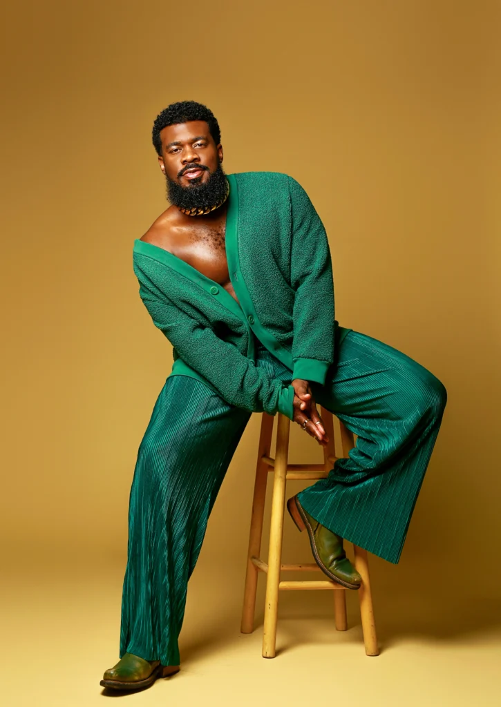 JaQuel Knight looks warmly at the camera. He leans to one side as he sits on a high stool. He wears a bright green cardigan, green satin trousers, and green leather shoes. The backdrop is a deep gold.