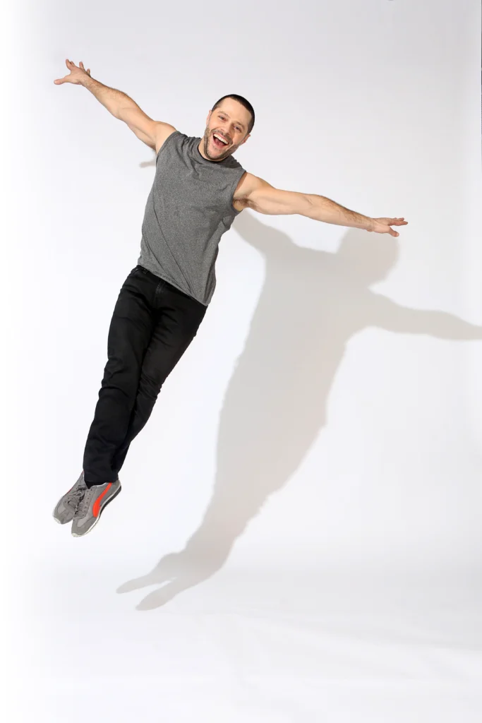 Joshua Bergasse grins widely at the camera as he is caught midair in an assemblé. He wears sneakers, black sweatpants, and a grey sleeveless shirt. His shadow dances on the white wall behind him.