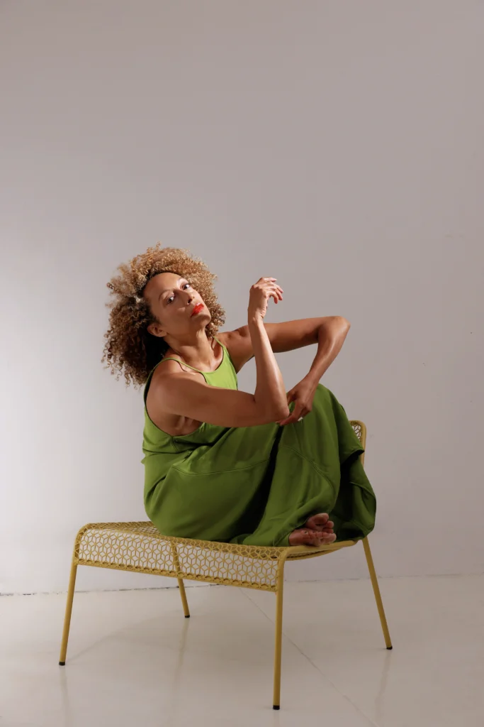 Stefanie Batten Bland sits on a yellow chair. Her knees are pulled in toward her chest. She tips her head back to gaze at the camera. Her arms are bent and angular, one hand crossing over her knees to cup the opposite elbow. Her brown curls are loose and halo out from her head. She wears red lipstick and a grass green jumpsuit.
