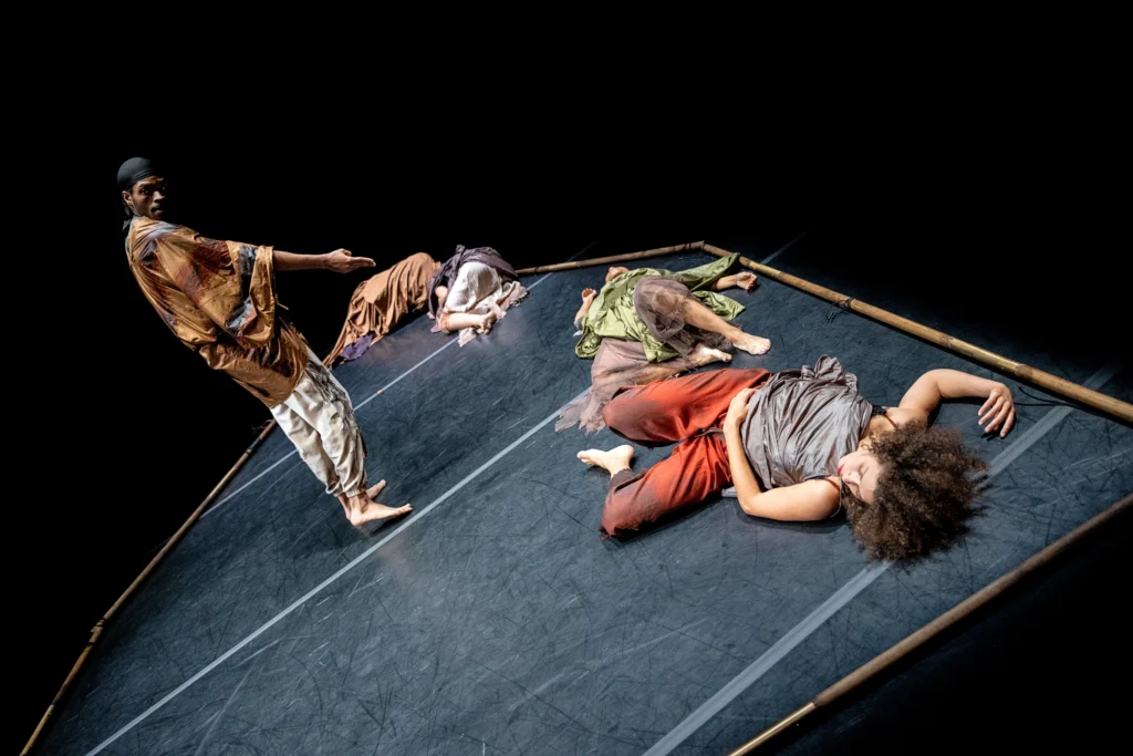 An off-kilter image that evokes a ship rocking on waves. Wooden sticks laid on the marley floor create the outline of a boat; the space beyond their borders is dark. Four dancers lie on their sides and backs as though exhausted. A fifth looks over his shoulder as he stands, gesturing down toward them.