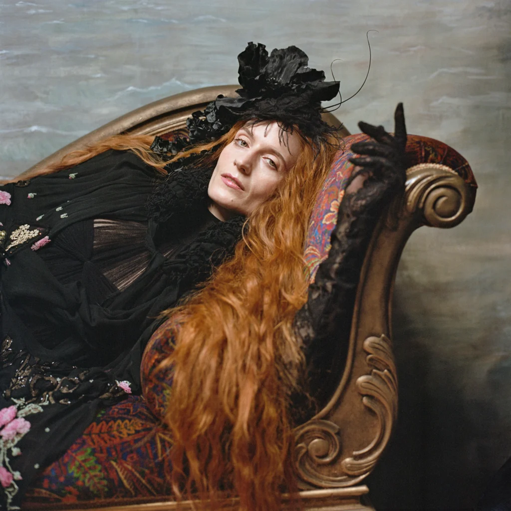 A woman reclines on a Victorian chaise lounge, gazing idly toward the camera. Long orange hair cascades over the side. She wears black lace and a matching fascinator.