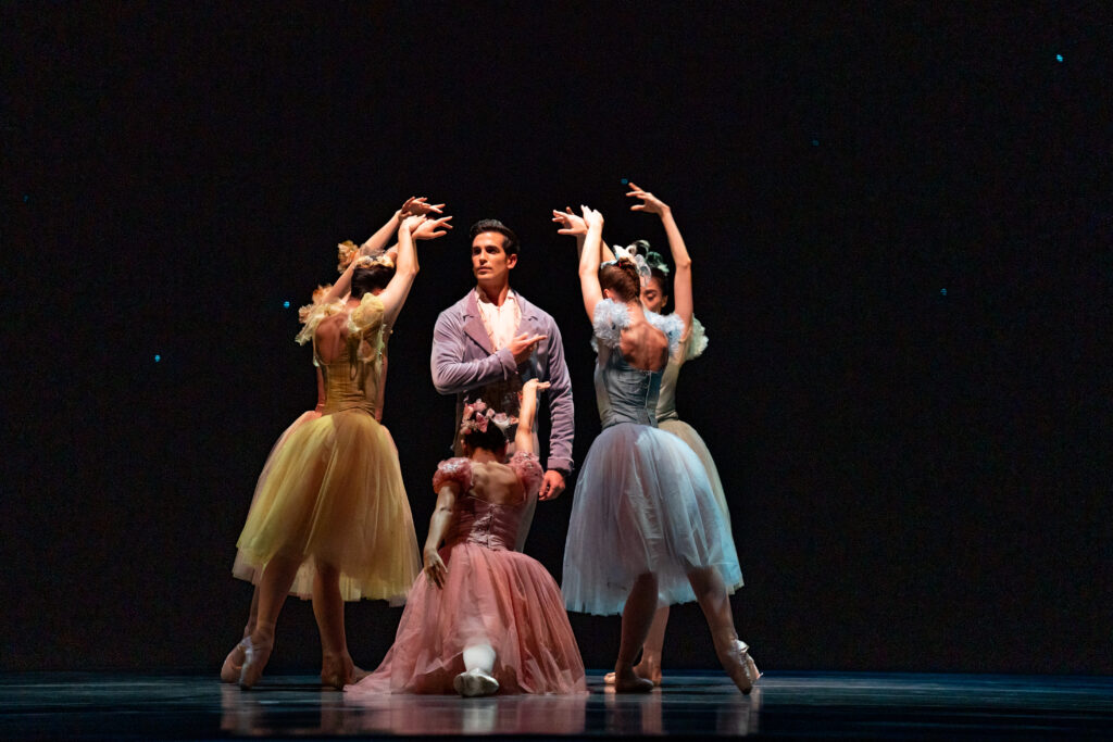 a male dancer surrounded by five female dancers with their arms reaching towards him and their heads bowed 
