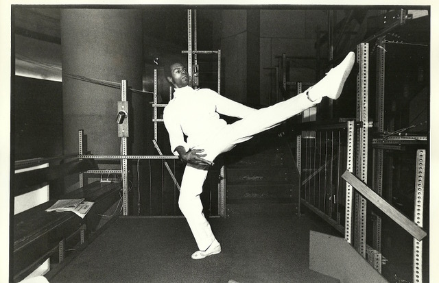 In a black and white archival photo, Gus extends a long leg to the side, twisting his torso towards it as his standing leg pliés. He seems larger than life amidst the pedestrian space; to one side is a bench, around him railings, behind him industrial steps.