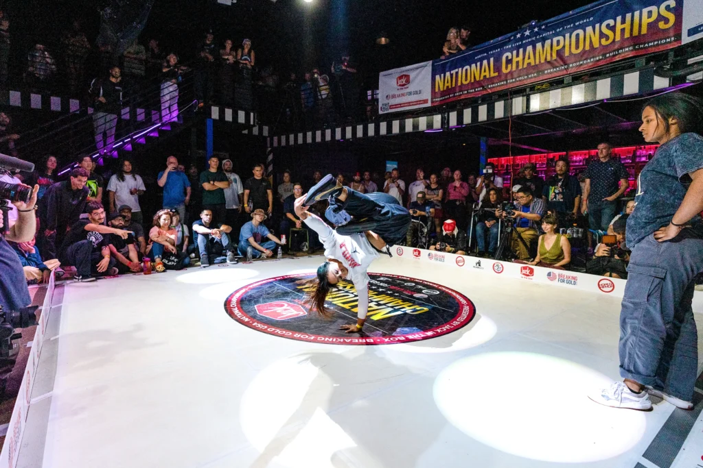 A breaker at the center of the floor balances on one hand, the other pulling a foot towards her head. A banner in the background reads "National Championships." Spectators sit and stand in layers around the floor.