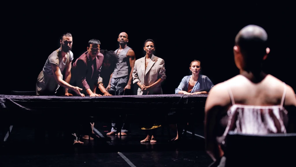 In the foreground to the right, a dancer sits in the chair, back to the camera as they look upstage. Six dancers stand or sit behind a table draped in black. One gestures to it expectantly, leaning forward; two others have their hands clasped before them, giving off a cold sense of welcome.