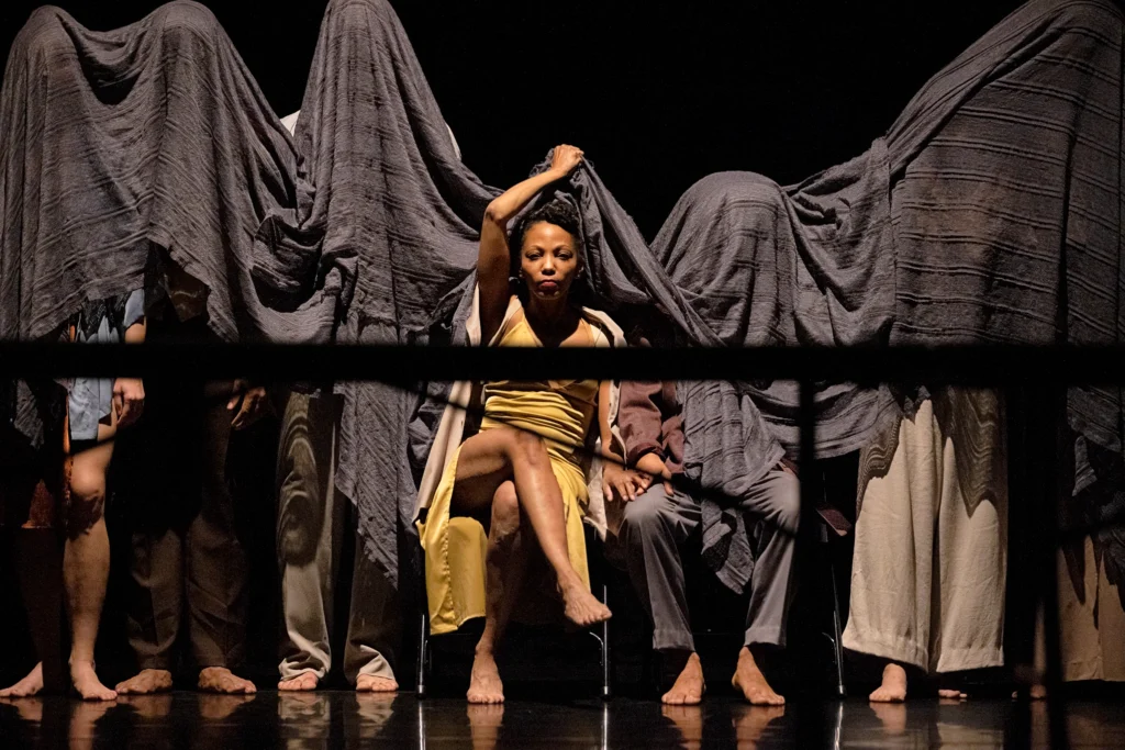 A woman in a yellow dress sits, legs crossed, behind a table that bisects the image. She holds a large, textured black cloth above her head with one hand, keeping it from covering her. To either side sit and stand other dancers, legs just visible as their upper bodies are hidden beneath the black cloth.