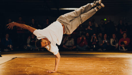 Morris "Bboy Morris" Isby balances on one arm, his legs both extended to 2 o'clock while his head is at 8 o'clock. He wears a white t-shirt, grey sweats, and well worn black Converse. Many members of the crowd watching at the side of a wooden dance floor have expressions of shock and delight.