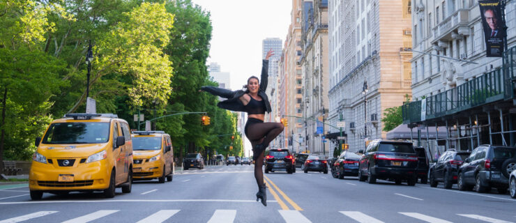 a dancer wearing black jumping in a parallel passe in the middle of a busy street