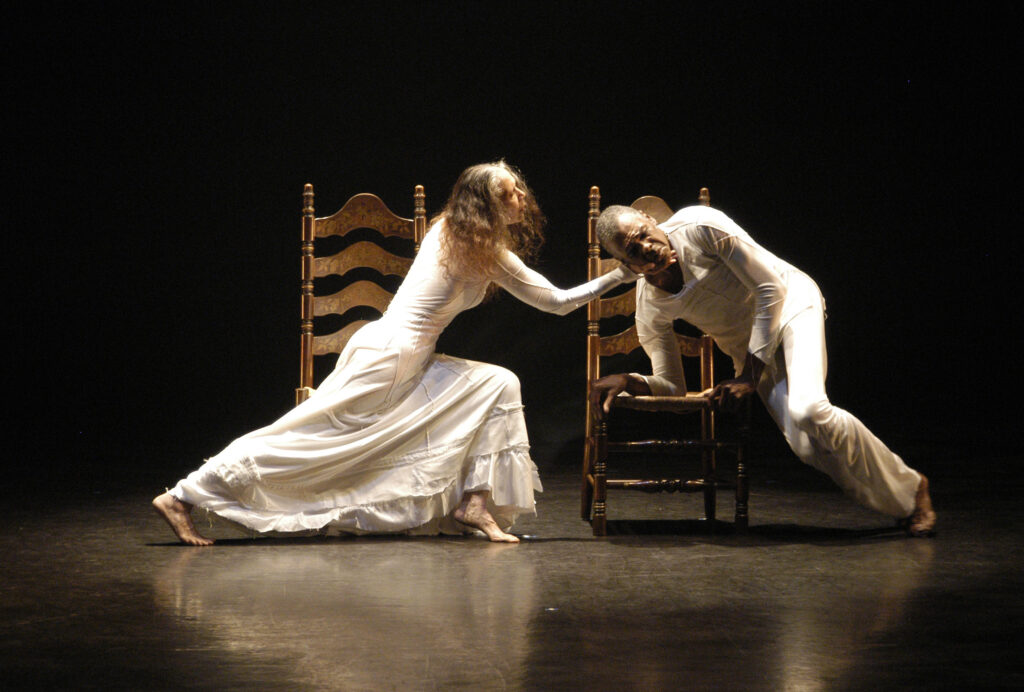 Carmen perches on a wooden chair, legs stretching into a lunge as she reaches to support Gus's head. Gus is beside an identical wooden chair, but he holds himself up with one hand planted on the seat, his knees bent as they perch on the ground to the side. Both wear white.