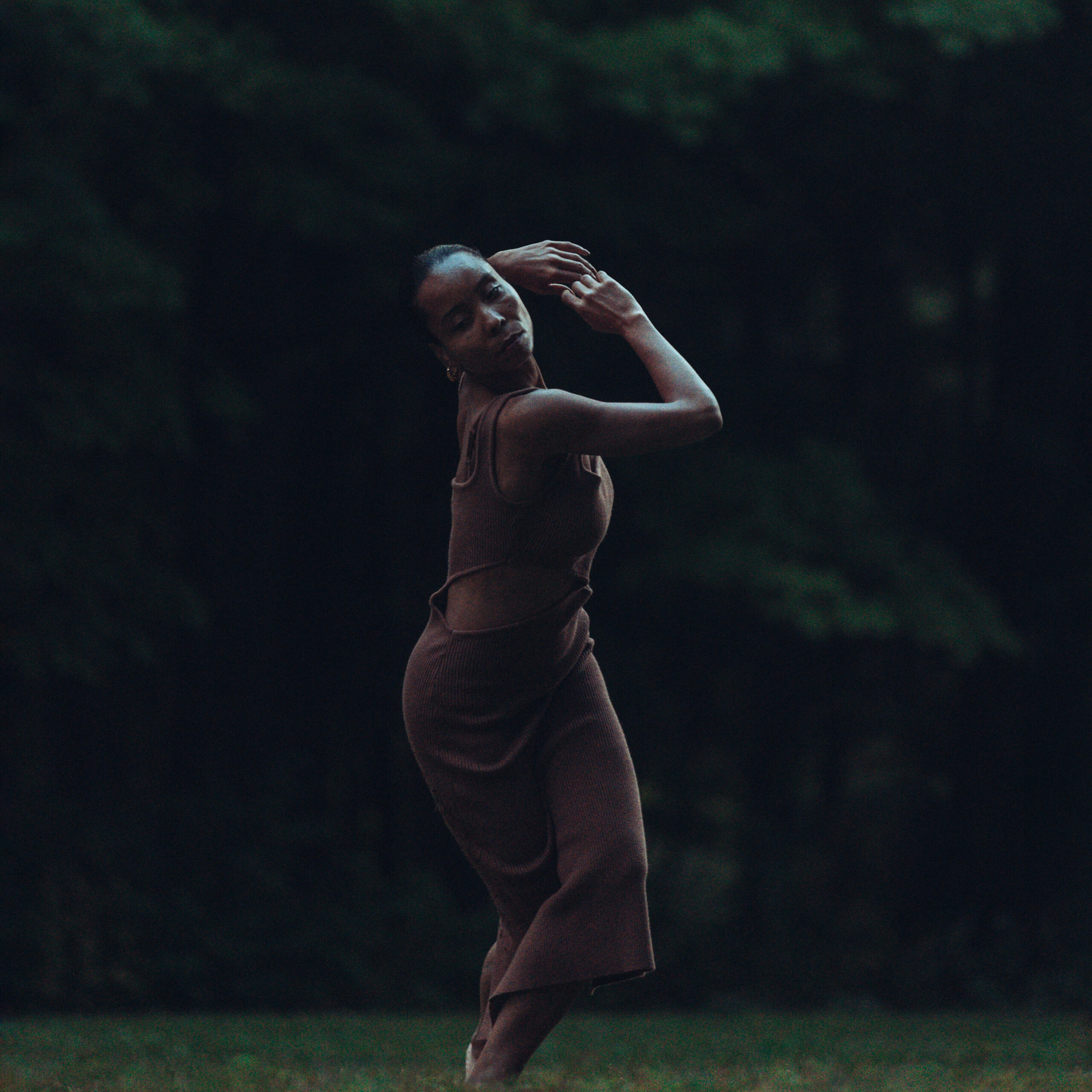 a female dancer standing in the grass, legs crossed and arms raised